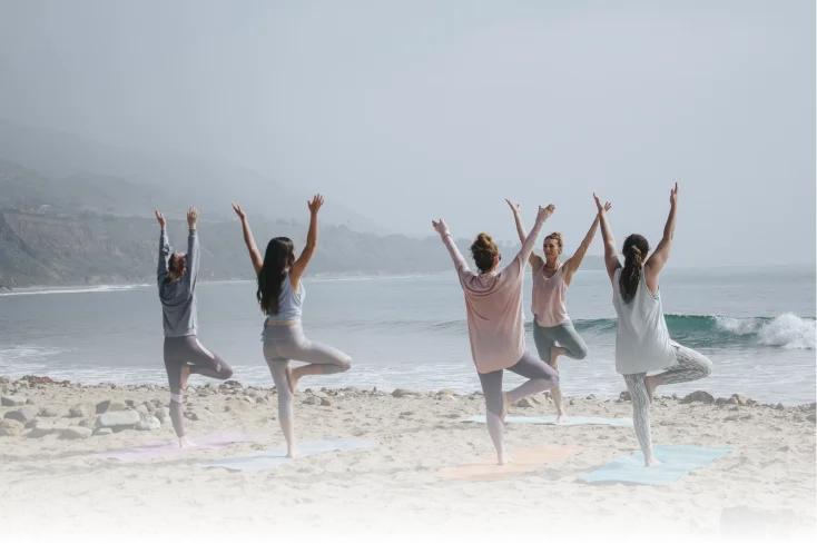 Five people practicing tree pose on yoga mats on a beach