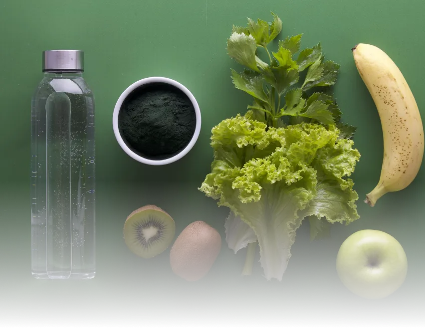 Water bottle, greens powder, kiwi, lettuce, celery, banana, and apple arranged on a green surface