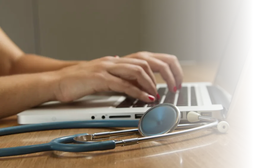 Hands typing on a laptop with a stethoscope on the desk